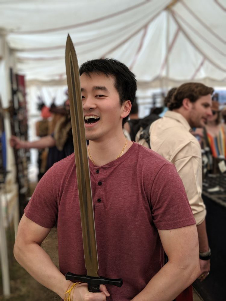 Don, a smiling Asian man, wearing a heathered maroon henley tee and yellow thread bracelets on his right wrist holds a golden sword with a black hilt in front of him.