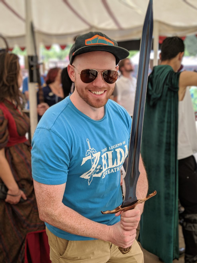 Mike, a smiling Caucasian man wearing aviator sunglasses, a light blue The Legend of Zelda Breath of the Wild t-shirt, tan pants, and a ball cap, holds a silver sword with a bronze hilt in front of him.