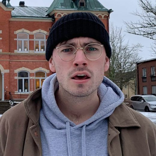 Photo of a man in glasses and a black beanie hat wearing a gray hoodie and tan jacket.
